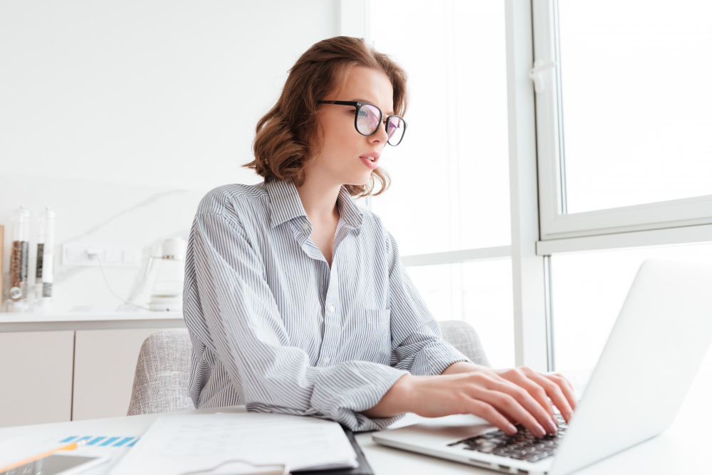 young-serious-woman-striped-shirt-typing-email-her-boss-while-sitting-workplace-light-apartment